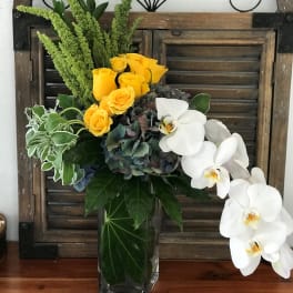 Yellow roses and white orchids in a glass vase with mixed greenery