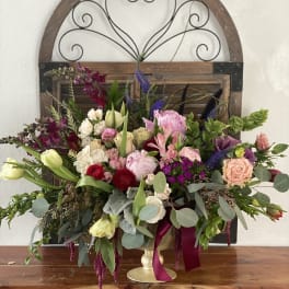 Large mixed flower arrangement in a gold pedestal vase with burgundy ribbon