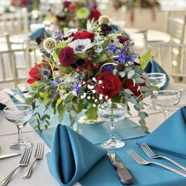 Low centerpiece with red roses, blue flowers, and greenery on a banquet table