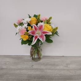 Mixed bouquet of pink lilies, yellow roses, white daisies, and carnations in a clear glass vase