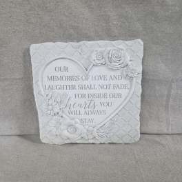 Square white memorial plaque with heart and sculpted flowers on a black metal stand.