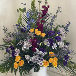 Tall mixed floral arrangement in a white pedestal vase with yellow, purple, and white blooms