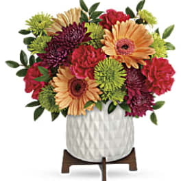 Compact bouquet of orange gerbera daisies, red carnations, and green mums in a white textured pot on a wooden stand