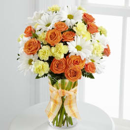 Orange roses, white daisies, and pale yellow carnations in a glass vase with a ribbon