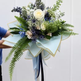 Handheld bouquet of white roses, blue flowers, and fern fronds wrapped in pale blue paper