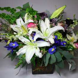 Mixed bouquet with white lilies, blue irises, and pink accents in a square vase