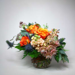 Orange roses arranged in a glass bowl with blue thistle and pale pink blooms