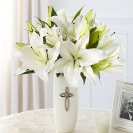White lilies arranged in a tall white vase with a cross emblem