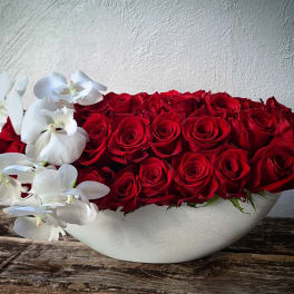 Red roses arranged in a white bowl with white orchids draping over the side