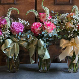 Three small glass vases with pink and peach roses, white fillers, and large satin ribbon bows.