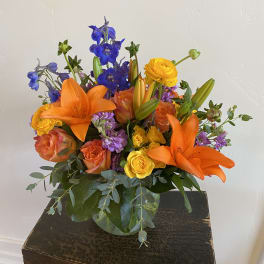 Bright mixed bouquet with orange lilies, roses, and blue flowers in a glass vase