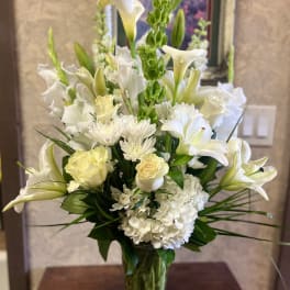 White floral arrangement in a clear glass vase with lilies, roses, and hydrangea