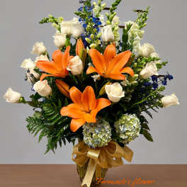 Tall arrangement of orange lilies, white roses, blue flowers, and hydrangeas in a gold vase with a bow