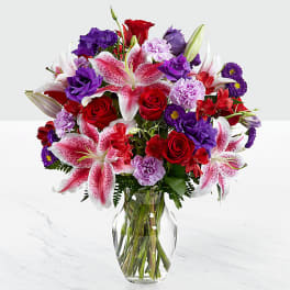 Bouquet of red roses, pink lilies, and purple flowers in a clear glass vase