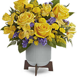 Yellow roses and yellow blooms with purple accents in a white ribbed pot on a wooden stand