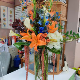 Tall mixed bouquet with orange lilies, blue accents, and white blooms in a glass vase
