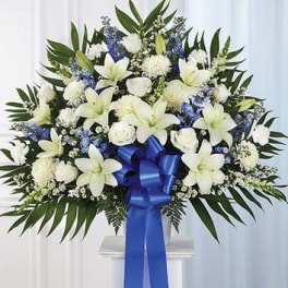 White lilies and roses with blue flowers in a standing spray with a blue ribbon