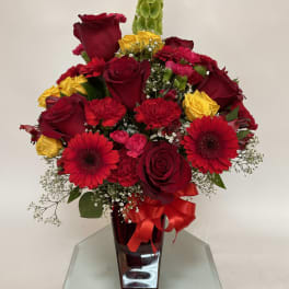 Red and yellow rose bouquet in a tall glass vase with a red ribbon