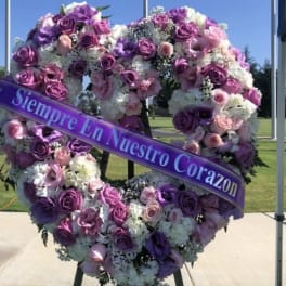 Heart-shaped floral wreath with purple and pink roses and a ribbon reading "Siempre En Nuestro Corazon"