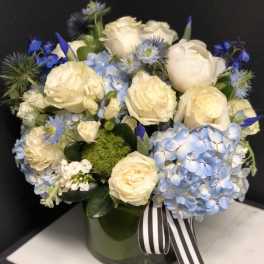 Cream roses and blue hydrangeas arranged in a glass vase with a striped ribbon.