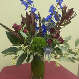 Tall vase arrangement with blue flowers, burgundy roses, and a green moss ball