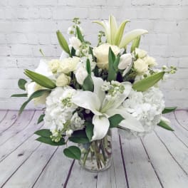White lilies, roses, hydrangeas and other white blooms arranged in a clear glass vase