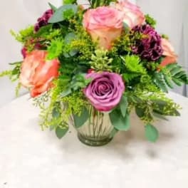 Round arrangement of peach and lavender roses with dark plum flowers in a clear glass vase