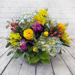Mixed bouquet of yellow roses, blue hydrangeas, and purple flowers in a glass vase