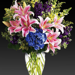 Pink lilies, blue hydrangeas, and purple flowers in a clear vase