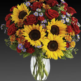 Bouquet of sunflowers, red roses, and small purple flowers in a clear vase