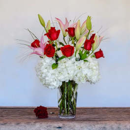 Tall vase arrangement of red roses, pink lilies, and white hydrangeas with one loose rose beside it.
