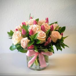 Blush roses and pink tulips arranged in a clear glass vase with a pink ribbon.