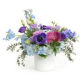 Low arrangement of blue hydrangea and purple blooms in a white cylinder vase