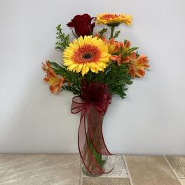 Tall clear vase with yellow gerbera daisies, orange alstroemeria, and a red rose with a burgundy ribbon bow.
