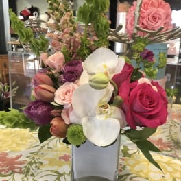 Mixed bouquet of pink roses, white orchids, and purple blooms in a square vase