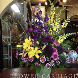 Tall mixed flower arrangement in a green vase with yellow lilies and purple blooms