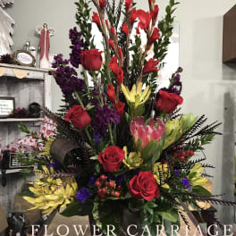 Tall mixed floral arrangement with red roses, purple blooms, and yellow flowers in a dark container