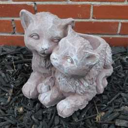 Two small cat figurines sitting together on mulch in front of a brick wall.