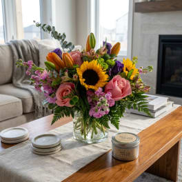 Mixed bouquet in a clear glass vase with sunflowers, roses, lilies, and purple blooms