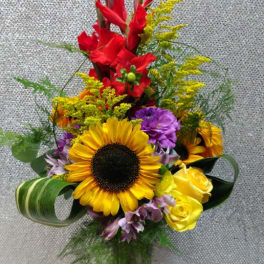Tall mixed bouquet with sunflowers, red gladiolus, and yellow roses in a clear vase
