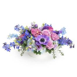 Low arrangement of purple anemones, lavender roses, and blue flowers in a white bowl.