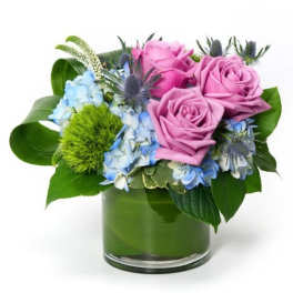 Short arrangement of pink roses, blue hydrangea, and green dianthus in a leaf-lined glass vase.