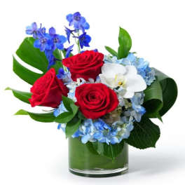 Short arrangement of red roses, blue flowers, blue hydrangea and a white orchid in a glass vase