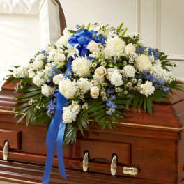 White and blue funeral casket spray with roses, lilies, and mums accented by a blue ribbon on a wood casket