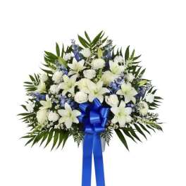 Standing spray of white lilies, roses, and carnations with blue flowers and a blue ribbon bow