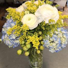 Bouquet of white ranunculus, blue hydrangea, and yellow filler in a glass vase
