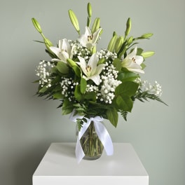 White lilies arranged in a glass vase with baby's breath and a white ribbon