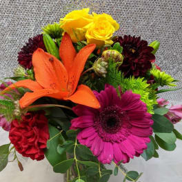Colorful bouquet with orange lily, pink gerbera, yellow roses, and red carnations in a glass vase