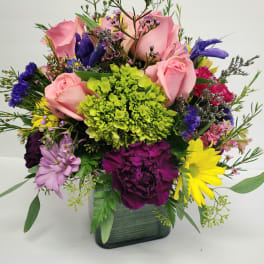Mixed bouquet of pink roses, daisies, and colorful filler flowers in a square vase