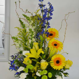 Tall mixed bouquet in a blue pot with yellow gerberas, blue delphinium, and white lilies